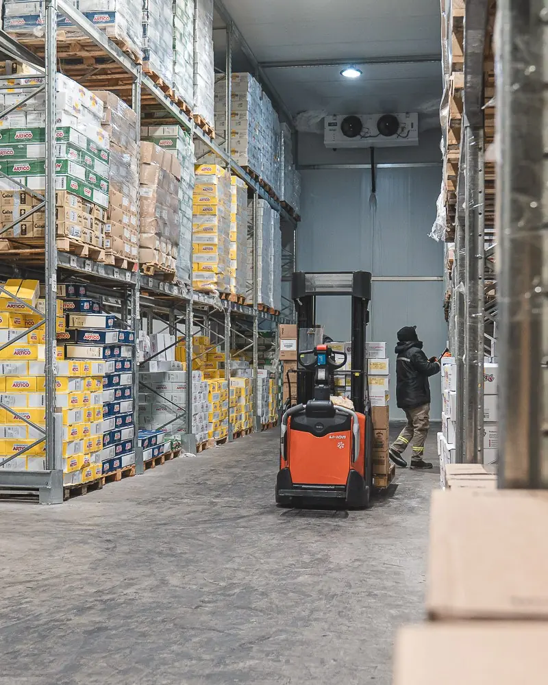 Un operario con ropa térmica y una zorra eléctrica naranja en el pasillo de la cámara de congelados, entre racks de cajas